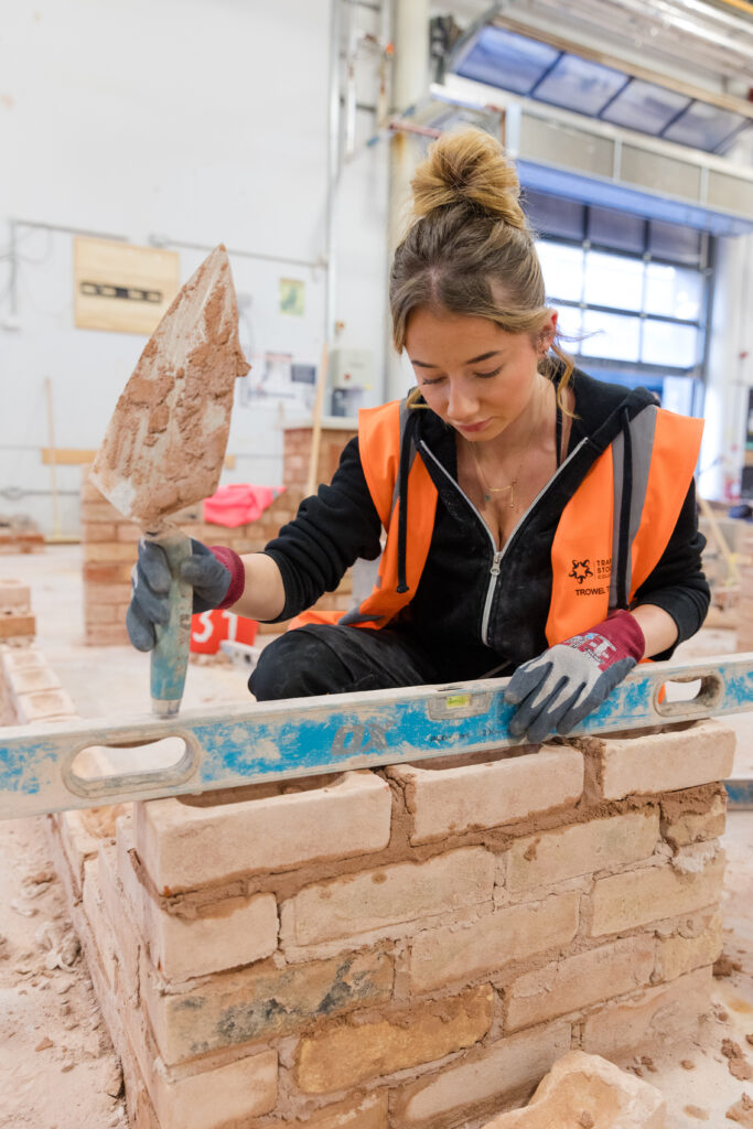 female student wearing PPE holding a trowel laying bricks building a wall