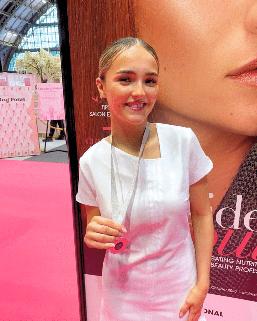 Smiling Amelia Yaqub in white uniform holding a medal in front of a Professional Beauty promotional backdrop.