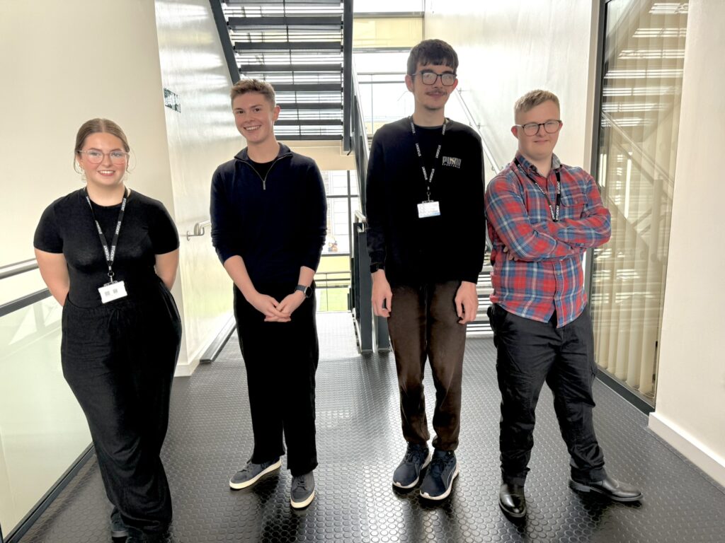 Four individuals wearing name badges standing side by side in a hallway with a staircase in the background