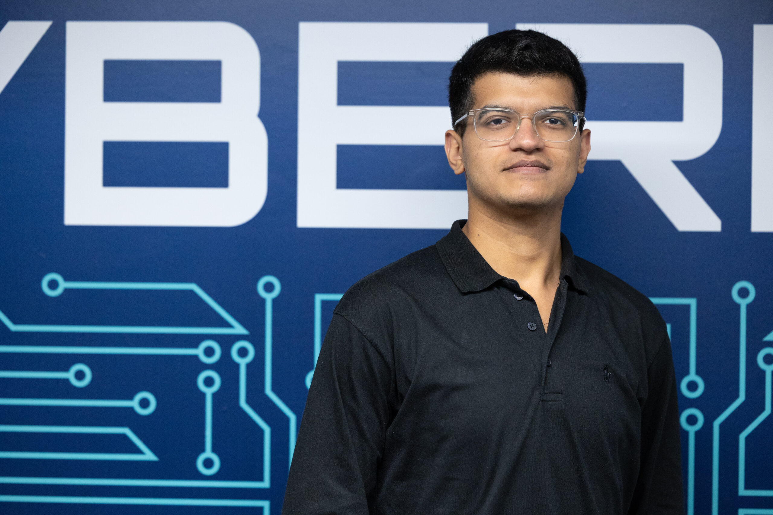 Hammad Jabeen standing in front of a blue wall with circuit board graphics and the word ‘CYBER’ visible, representing a cybersecurity learning environment